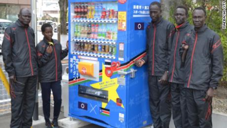 The South Sudan team and their coach, Joseph, (far left) in front of a vending machine in Maebashi city, displaying their nation&#39;s flag. 